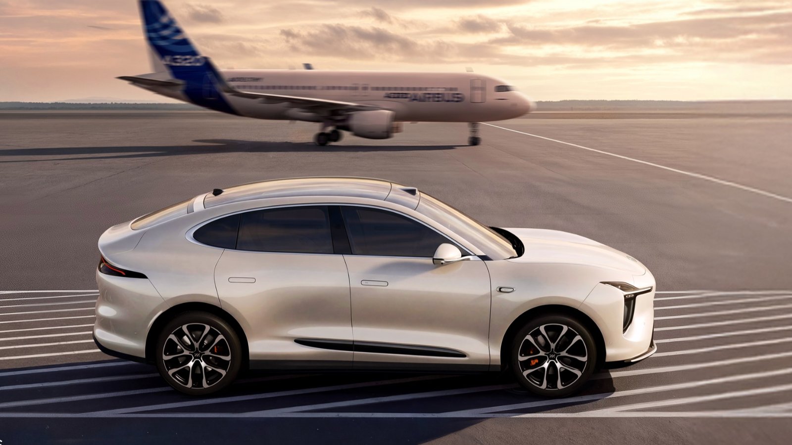 Side profile of a silver IM Motors LS6 on an airport apron with a passenger aircraft in the background.